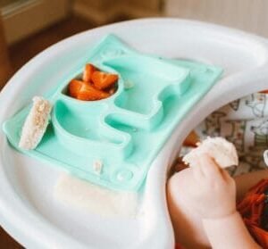 Tanjir u obliku slona sa hranom za bebe na hranilici i bebinom rukom koja uzima hranu.